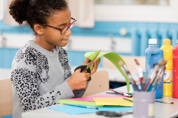 Kind schneidet buntes Papier im Kunstunterricht Ein Mädchen sitzt im Klassenraum am Tisch, schneidet grünes Papier mit einer Schere und ist von Bastelmaterialien, Pinseln und bunten Farben umgeben.